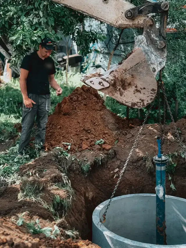 Worker watching an excavator set a concrete tank in a backyard trench